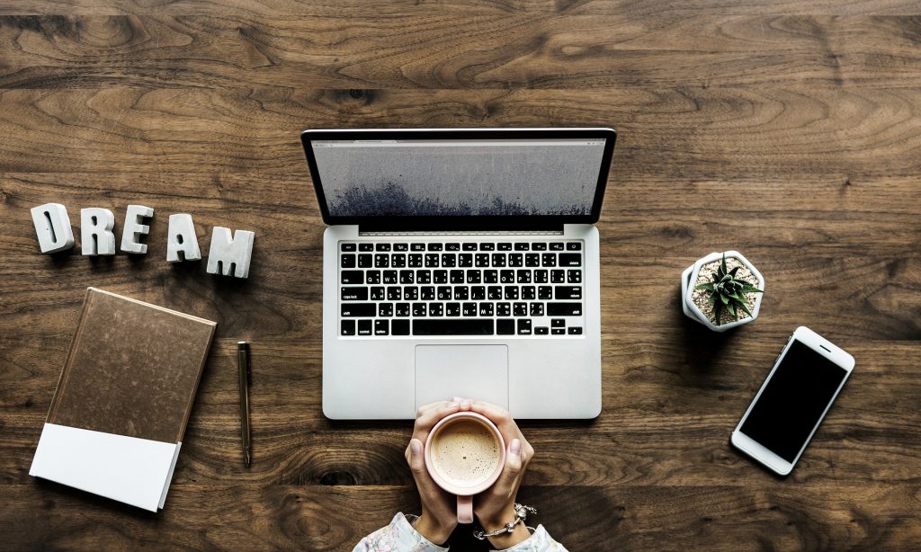 A brown desk where a student sits grasping a coffee mug in front of a lap top with a brown notebook, pencil, cell phone, small green plant and a DREAM sign