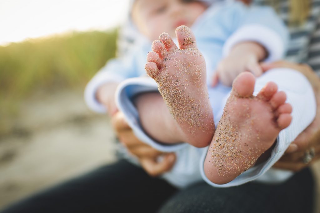 Closeup of infant's bare feet and toes with sand on them.  Infant is wearing light blue and held by parent.