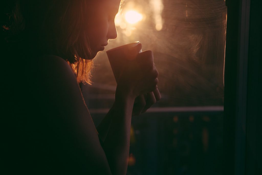 Profile of sad girl looking down with eyes closed holding coffee cup next to a window with sunlight streaming through.