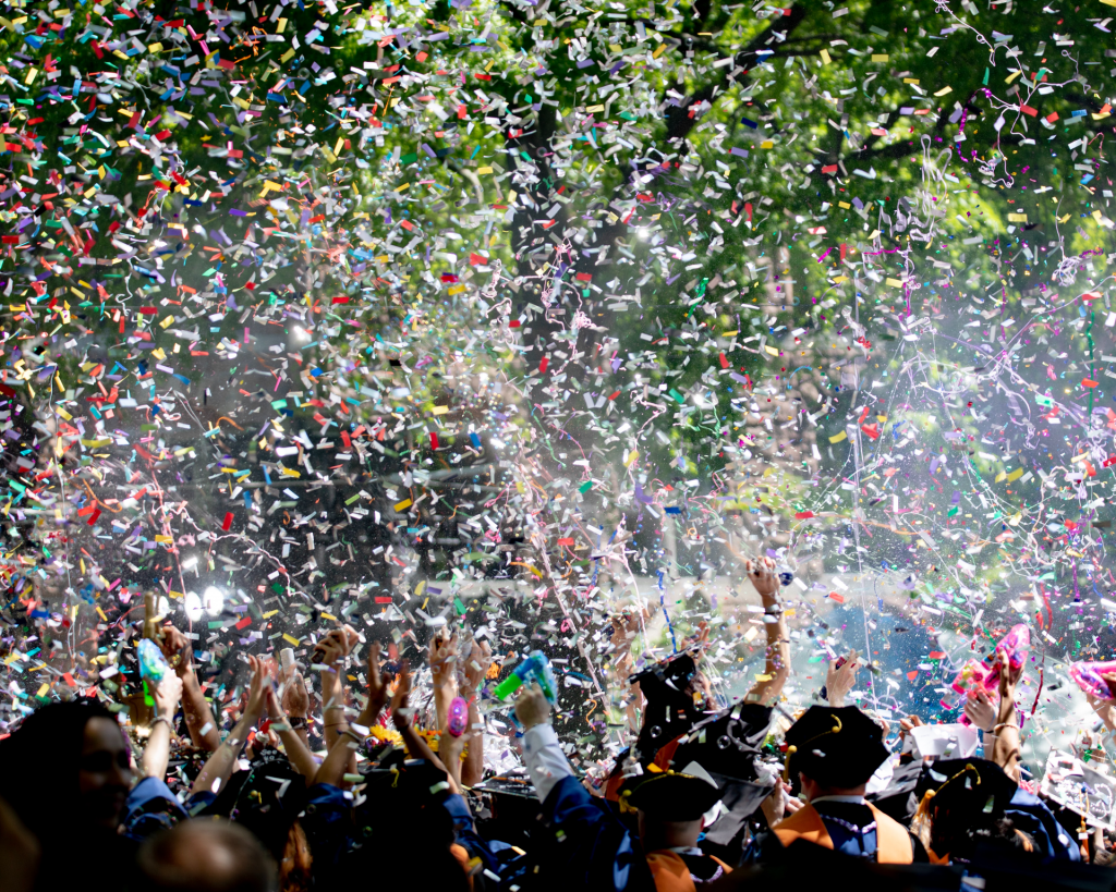 Graduating class celebration with graduates' arms in air with multi-colored confetti flying in air.