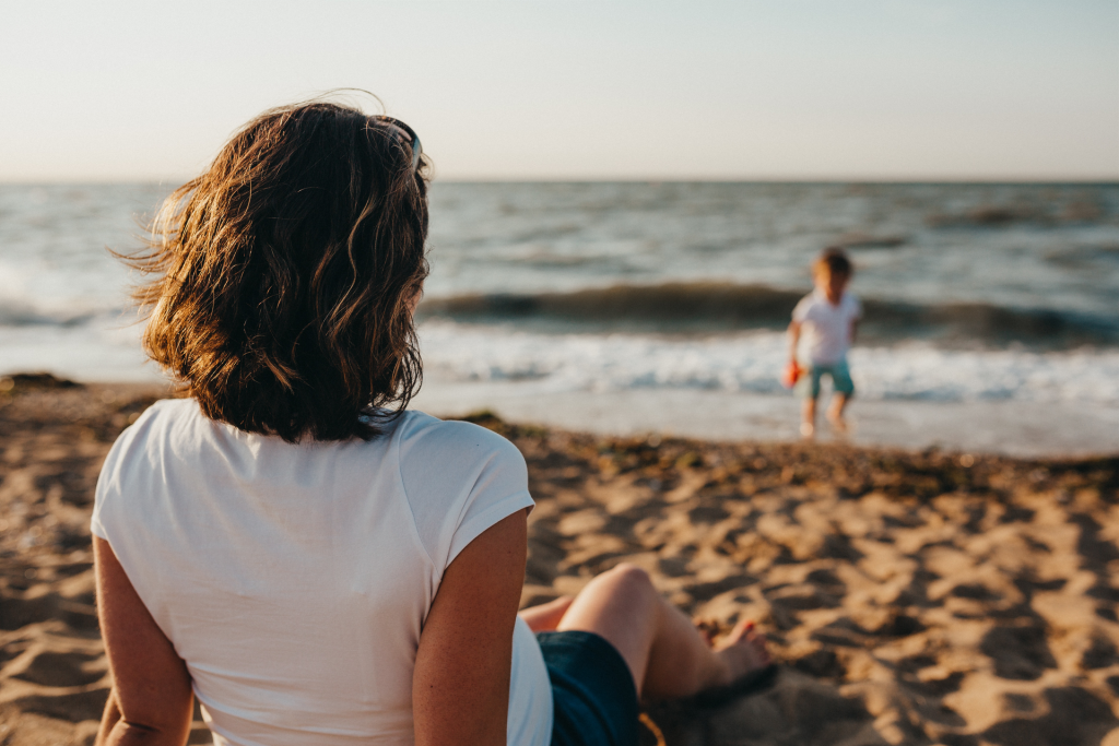 Mom sitting on beach facing ocean and watching small child playing on beach in sunlight