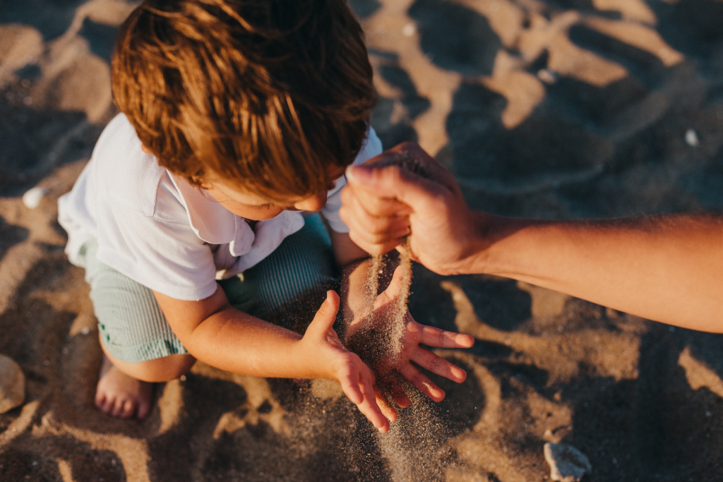 Toddler boy crouched on beach holding hands open to catch sand coming from mother's hand