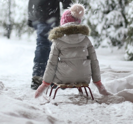 Little girl sitting on sled in snow wearing a pink beanie hat with light pink pom pom