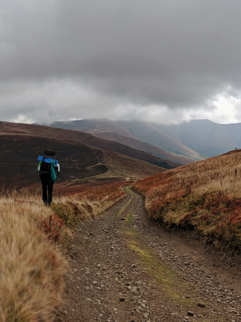 Hiker on desolate gravel road heading towards mountains and grey skies.