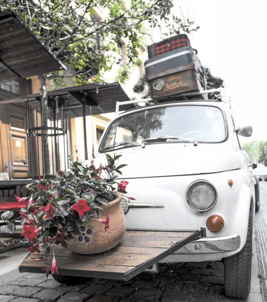 White car with multiple suitcases on top and a flower pot hanging on the front end