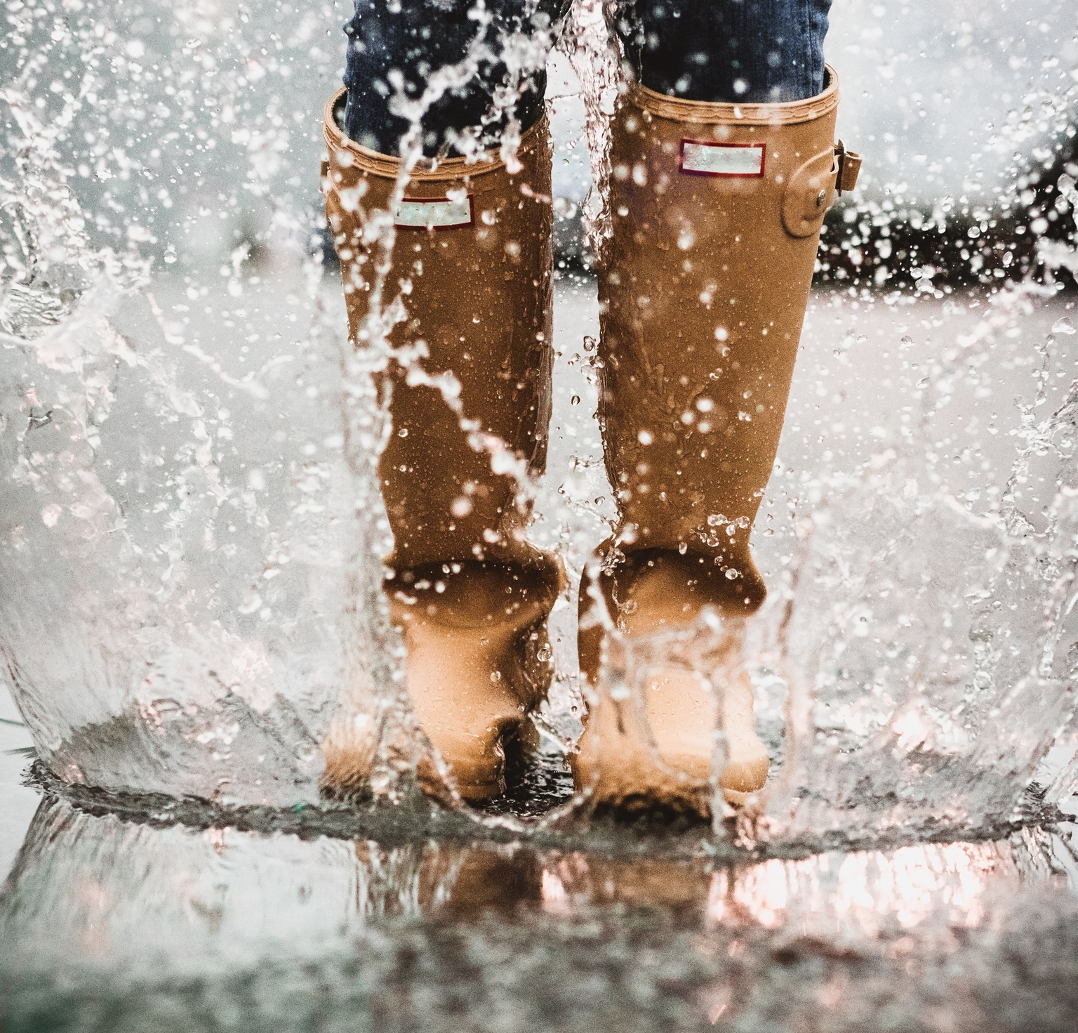 Close up of woman wearing tan colored rain boots splashing in a puddle of water 
