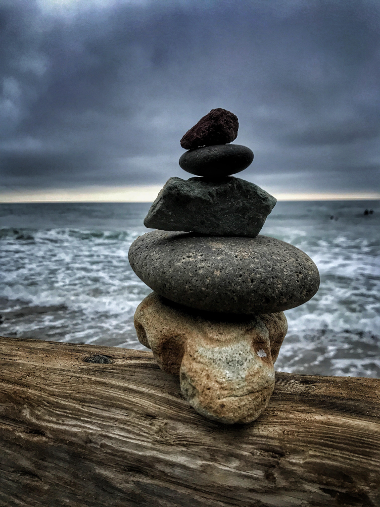 Five beach rocks stacked on top of one another on driftwood with sea in background