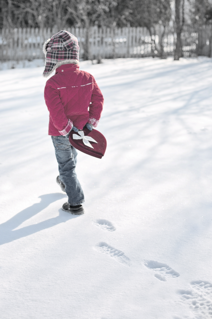 Small boy, wearing red winter jacket, carrying a red valentine box behind his back walking in snow