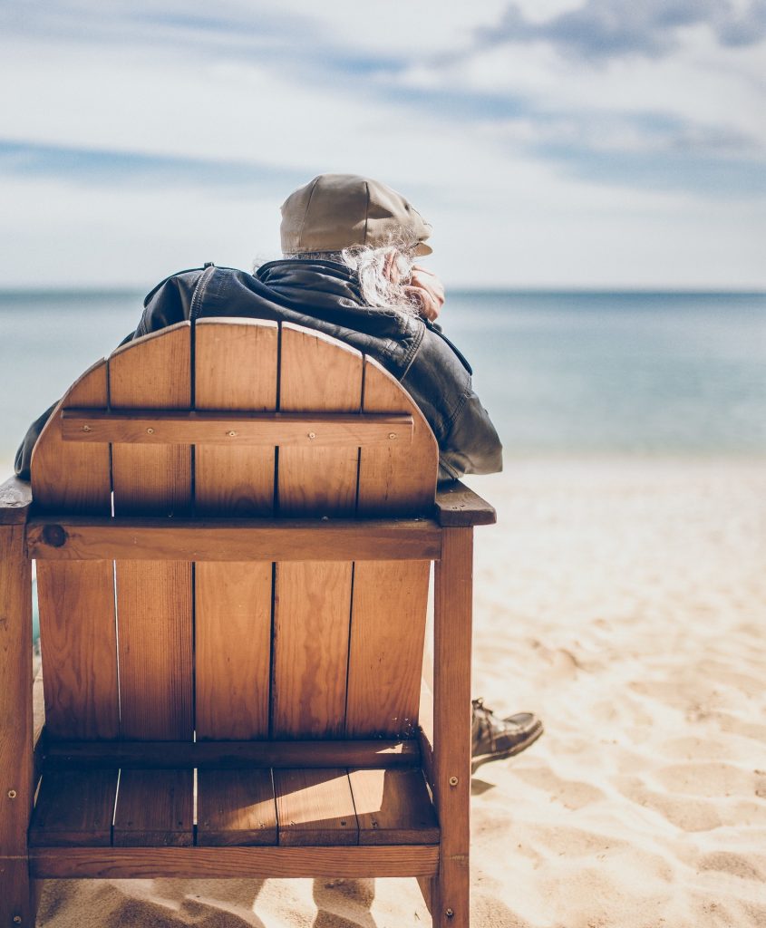 Older man sitting in wood chair on beach looking at ocean